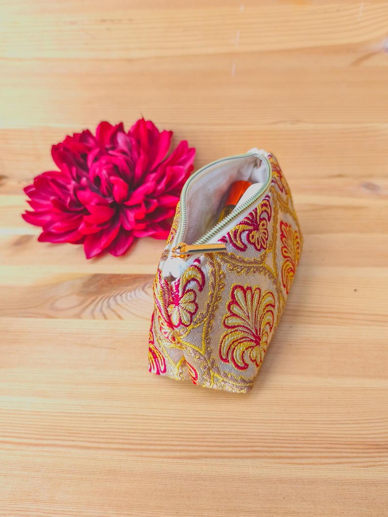 May include: A beige cosmetic bag with a gold zipper, embroidered with red and yellow floral patterns. A large, vibrant pink flower sits beside the open bag. The bag contains a small orange bottle.