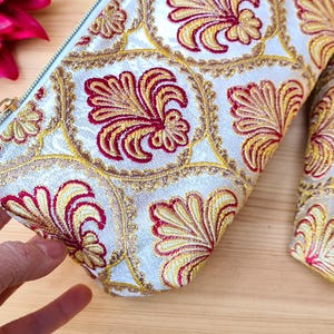 May include: A white cosmetic bag with a repeating pattern of red and yellow floral designs. The bag has a light blue zipper and is made of a textured fabric. The bag is on a wooden surface.