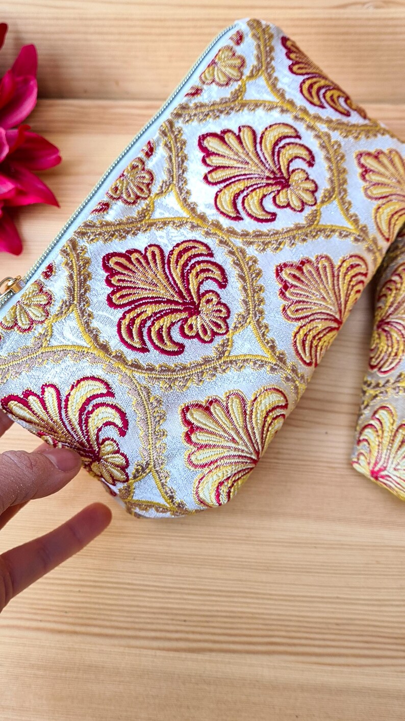 May include: A white cosmetic bag with a repeating pattern of red and yellow floral designs. The bag has a light blue zipper and is made of a textured fabric. The bag is on a wooden surface.