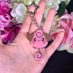 May include: Pink and brown glitter resin keychain featuring a cartoon character with a curled tail. The keychain has a silver star charm.