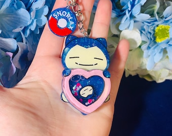 May include: A blue Snorlax charm with glitter embedded in the resin. The charm is shaped like a heart and has a pink border. The charm is attached to a silver key chain with a red and blue Snorlax charm.