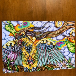 May include: A colorful, detailed drawing of a mythical creature with wings, multiple eyes, and a beak. The creature is surrounded by clouds, flowers, and leaves. The artwork is done in a vibrant style with a mix of blue, green, yellow, pink, and orange colors.