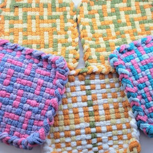 May include: Four woven pot holders in different colors and patterns. The pot holders are square and have a woven texture. The colors are pink, blue, green, and yellow.