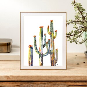 May include: A watercolor painting of a group of cacti with orange flowers. The cacti are painted in shades of green, blue, and yellow. The painting is framed in a gold frame.