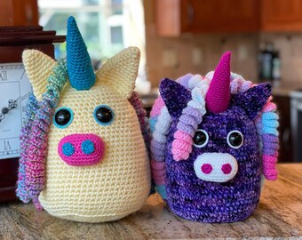 May include: Two crocheted unicorn plushies, one yellow with a blue horn and the other purple with a pink horn. Both have pink noses and large black eyes.