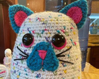 May include: A white crocheted cat with colourful speckles, large black eyes, a pink nose, and a teal blue tail and ears.