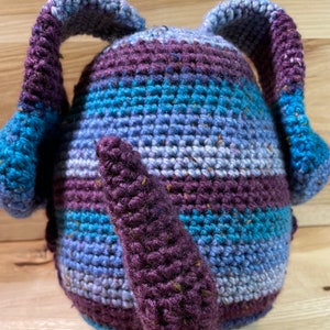 May include: A crocheted stuffed toy with a purple tail and blue and purple striped body. The toy has two long, floppy ears.