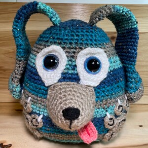 May include: A crocheted blue and brown dog toy with white eyes and a pink tongue. The toy has a textured surface and is made of yarn.