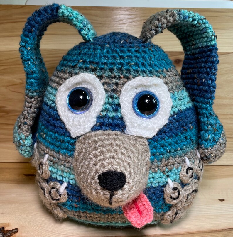 May include: A crocheted blue and brown dog toy with white eyes and a pink tongue. The toy has a textured surface and is made of yarn.