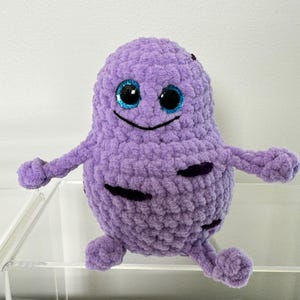 Puede incluir: Un juguete de peluche morado de crochet con forma de célula con detalles negros y ojos azules con purpurina. El juguete tiene una cara sonriente y dos brazos.