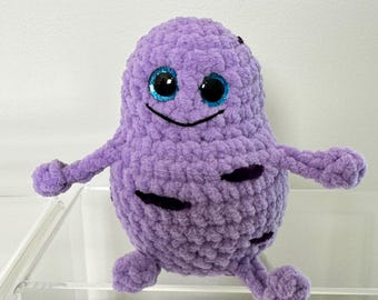Könnte beinhalten: Ein lilafarbenes Häkeltier in Form einer Zelle mit schwarzen Details und blauen Glitzeraugen. Das Spielzeug hat ein lächelndes Gesicht und zwei Arme.