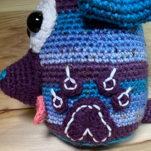 May include: A crocheted stuffed animal in the shape of a dog. The dog is blue, purple, and white with a pink tongue. The dog has a white paw print on its side.