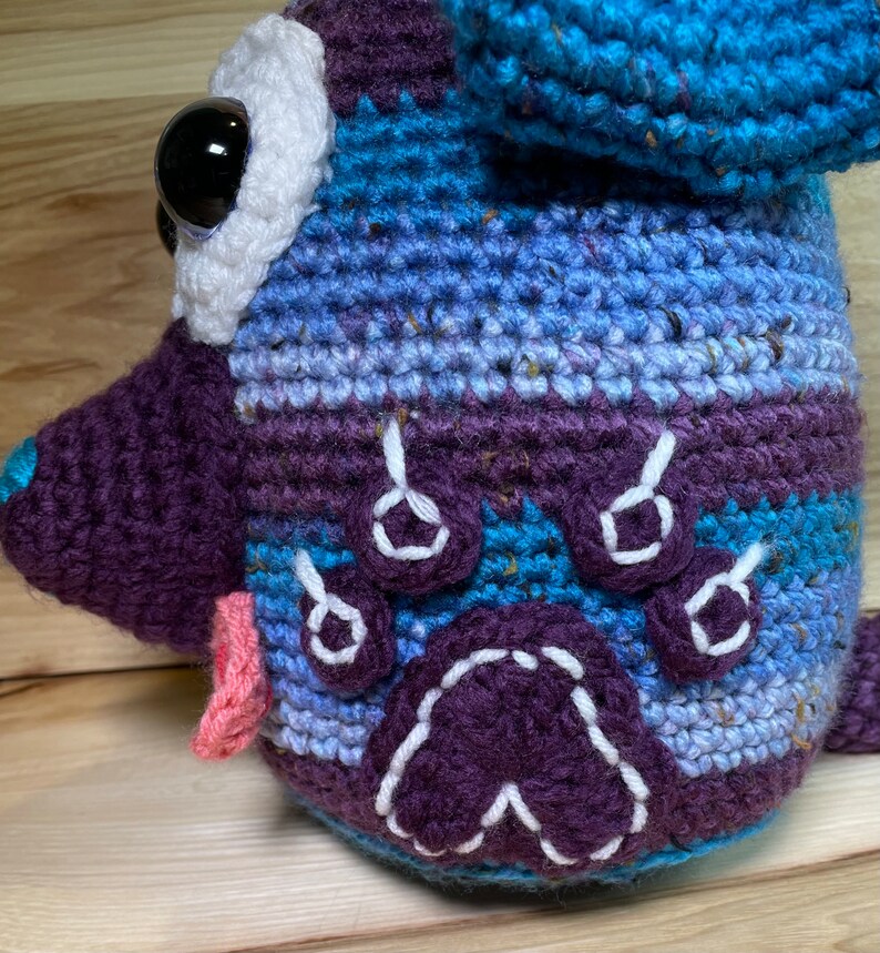 May include: A crocheted stuffed animal in the shape of a dog. The dog is blue, purple, and white with a pink tongue. The dog has a white paw print on its side.