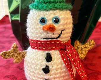 Könnte beinhalten: Ein gehäkelter Schneemann mit einem grünen Hut, einem rot-goldenen Schal und schwarzen Knöpfen. Der Schneemann hat eine Karottennase und blaue Augen.