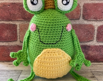 May include: A green crocheted frog wearing a gold crown with large white eyes and pink cheeks. The frog has a yellow crocheted belly and is sitting on a wooden surface.