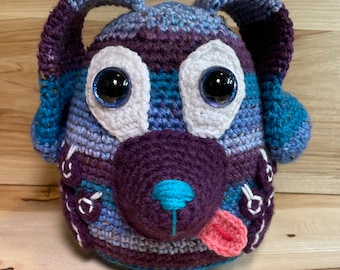 Könnte beinhalten: Ein gehäkeltes Stofftier in Form eines Hundes aus blauem, violettem und weißem Garn. Der Hund hat große, schwarze Augen mit weißen Pupillen und eine herausgestreckte rosa Zunge.