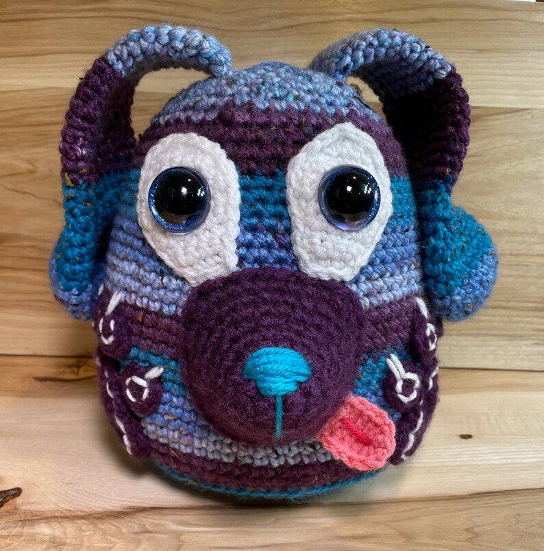 May include: A crocheted stuffed dog toy made with blue, purple and white yarn. The dog has large, black eyes with white pupils and a pink tongue sticking out.