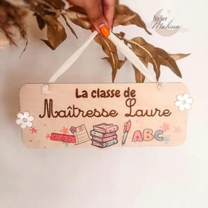 Peut inclure: Panneau de classe en bois avec le texte "La classe de Maîtresse Laure" et des illustrations de fournitures scolaires. Le panneau est rectangulaire avec un ruban blanc pour l'accrocher et des fleurs décoratives.