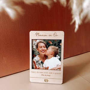 Peut inclure: Un cadre photo rectangulaire en bois clair présentant une photographie imprimée d'un enfant embrassant un adulte sur la joue. Le cadre porte l'inscription "Nounou en Or" en haut et "Merci de nous avoir aidé à grandir Alba & Hugo" en bas.