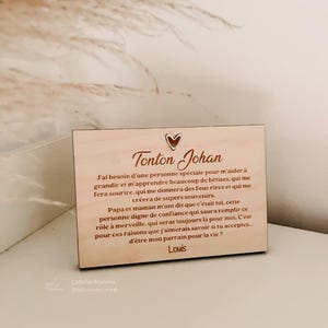 May include: A light brown wooden plaque with the words "Tonton Johan" and a heart symbol. The plaque has a message in French asking someone to be a godfather. The text is in a handwritten style. The plaque is rectangular.