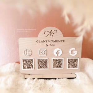 Peut inclure: Un présentoir en bois avec le texte "GLANZMOMENTE by Maria" et des icônes de réseaux sociaux. Le présentoir comporte des codes QR sous les icônes Instagram, Facebook et Google. Le fond est rose clair avec une surface blanche duveteuse.