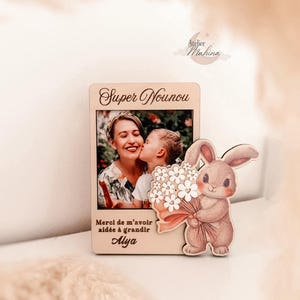 May include: A wooden photo frame featuring a photograph of a woman and child. The frame is inscribed with the words "Super Nounou" and "Merci de m'avoir aidée à grandir Alya". A cartoon bunny holding flowers is attached to the frame.
