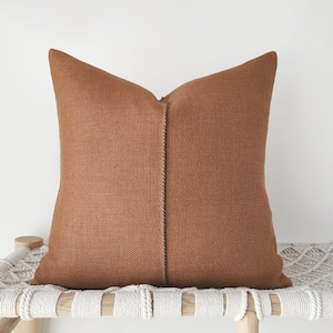 May include: A square throw pillow in a solid burnt orange color with a visible woven texture. The pillow features a vertical seam running down the center, creating a distinct line that divides the front surface into two symmetrical halves.