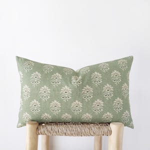 May include: A rectangular sage green pillow with a repeating floral pattern in beige and light gray. The pillow is resting on a light-colored wooden stool with a woven seat. The pillow's design adds a touch of elegance.