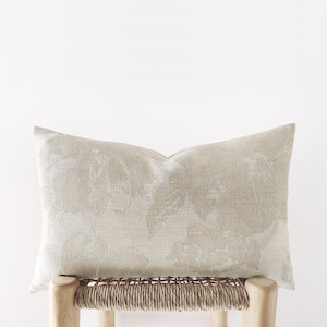 May include: A rectangular beige decorative pillow with a floral pattern. The pillow is placed on a light-colored wooden stool with a woven seat. The floral design is a lighter shade of beige, creating a subtle contrast.