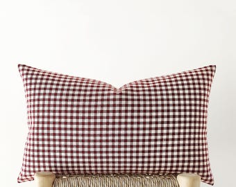 May include: A rectangular throw pillow featuring a classic dark red and white gingham check pattern. The pillow has a structured, plump appearance with a distinct center crease along the top edge, showcasing a woven fabric texture throughout.