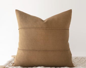 May include: A square throw pillow in a solid medium brown color. The pillow features a textured fabric surface with two horizontal stitched lines running across the center, creating a subtle layered appearance on the front face.