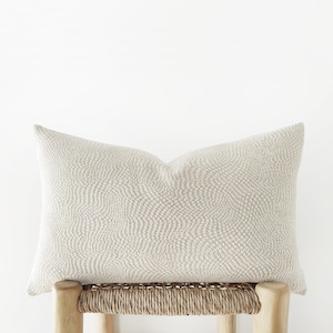 Op de afbeelding: Een rechthoekig decoratief kussen met een getextureerd, lichtbeige patroon. Het kussen ligt op een houten kruk met een geweven zitting. De neutrale kleur en het ontwerp suggereren een focus op interieurdecoratie.