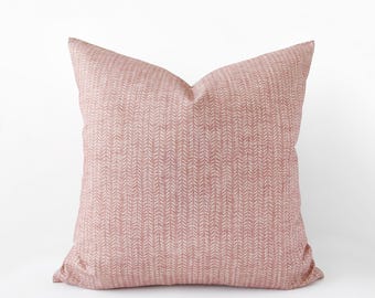 Puede incluir: Un cojín cuadrado con una base de tela en color rosa empolvado o terracota apagado. La superficie presenta un patrón vertical repetitivo de pequeñas líneas blancas discontinuas en forma de espiga o chevron, creando una textura sutil.