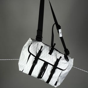 May include: White and black messenger bag with a diamond pattern. The bag has black straps and buckles, and a long shoulder strap. The bag is suspended in the air against a gray background.