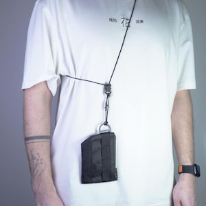 Peut inclure: Portefeuille tactique noir avec cordon, porté autour du cou. Le portefeuille est rectangulaire avec un anneau métallique et des sangles verticales. Le cordon noir se connecte au portefeuille avec un fermoir noir. Le t-shirt blanc porte des caractères chinois.