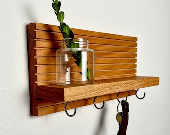 May include: A wooden wall shelf with a horizontal slat back and a lower shelf with three metal hooks. A clear glass jar with green leaves sits on the shelf. A set of keys hangs from one of the hooks. The wood has a warm, natural finish.
