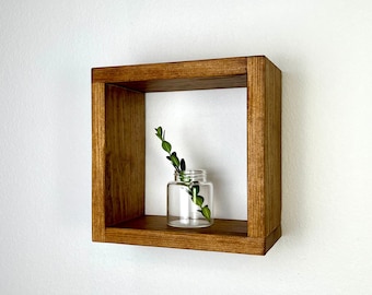 May include: A small, square wooden shelf with a single, clear glass jar containing a small sprig of greenery. The shelf is stained a dark brown color and is mounted on a white wall.