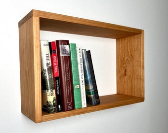 May include: A rectangular, light-brown wooden floating shelf with several books inside. The books have various colors and titles, including "The Toro", "not a fan.", and "The Millionaire Next Door". The shelf is mounted on a white wall.