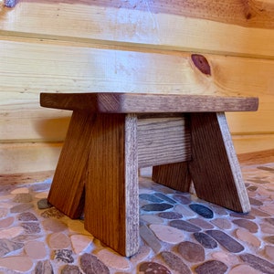May include: A small wooden stool with a natural brown finish. The stool has a rectangular top and three legs. The stool is sitting on a floor made of small, round stones.