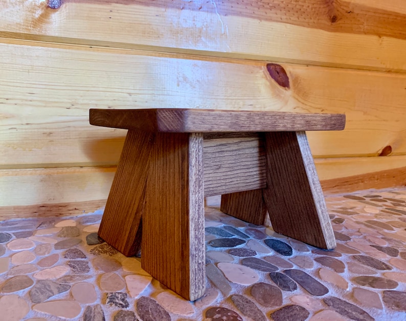May include: A small wooden stool with a natural brown finish. The stool has a rectangular top and three legs. The stool is sitting on a floor made of small, round stones.