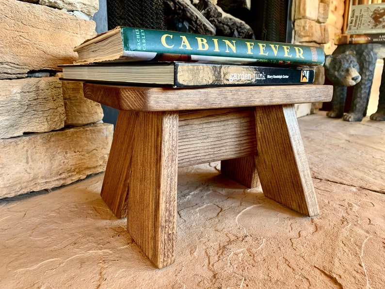 May include: A small wooden stool with a natural brown finish. The stool has a rectangular top and four legs. Three books are stacked on top of the stool, including one titled "Cabin Fever".