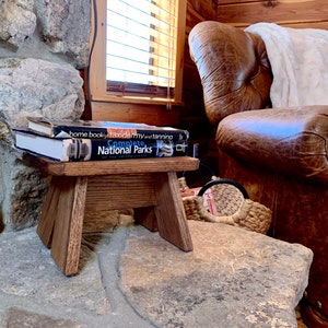 May include: A small wooden stool with a dark brown finish sits on a stone surface. Two books are stacked on top of the stool, one titled "Complete National Parks". A leather armchair with a white throw blanket is in the background.