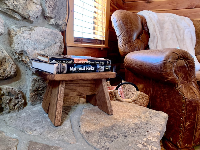 May include: A small wooden stool with a dark brown finish sits on a stone surface. Two books are stacked on top of the stool, one titled "Complete National Parks". A leather armchair with a white throw blanket is in the background.