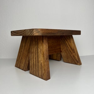 May include: A small wooden stool with a rectangular top and four legs. The stool is made of dark brown wood with a natural wood grain.
