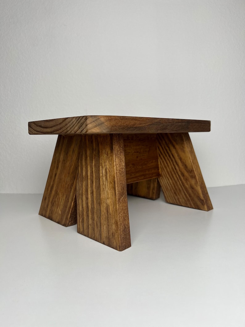 May include: A small wooden stool with a rectangular top and four legs. The stool is made of dark brown wood with a natural wood grain.