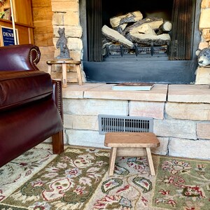 May include: A rustic wooden stool with a natural finish sits in front of a fireplace with a stone surround. The stool is in front of a patterned rug with shades of green, red, and beige.