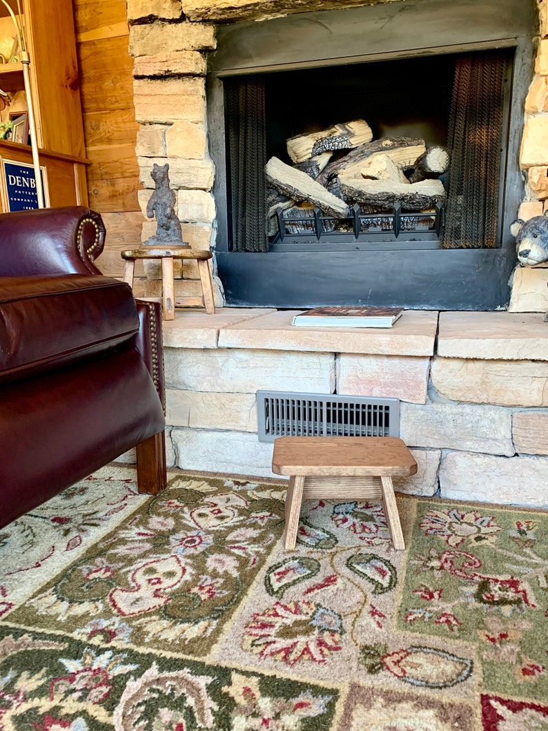 May include: A rustic wooden stool with a natural finish sits in front of a fireplace with a stone surround. The stool is in front of a patterned rug with shades of green, red, and beige.