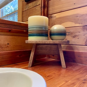 May include: A small wooden stool with two candles on top. The candles are striped with shades of blue, green, and brown. The stool is in a bathroom with wood paneling.