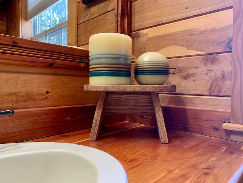 May include: A small wooden stool with two candles on top. The candles are striped with shades of blue, green, and brown. The stool is in a bathroom with wood paneling.