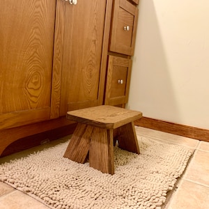 May include: A wooden step stool with a natural brown finish. The stool is sitting on a white shag rug in a bathroom.